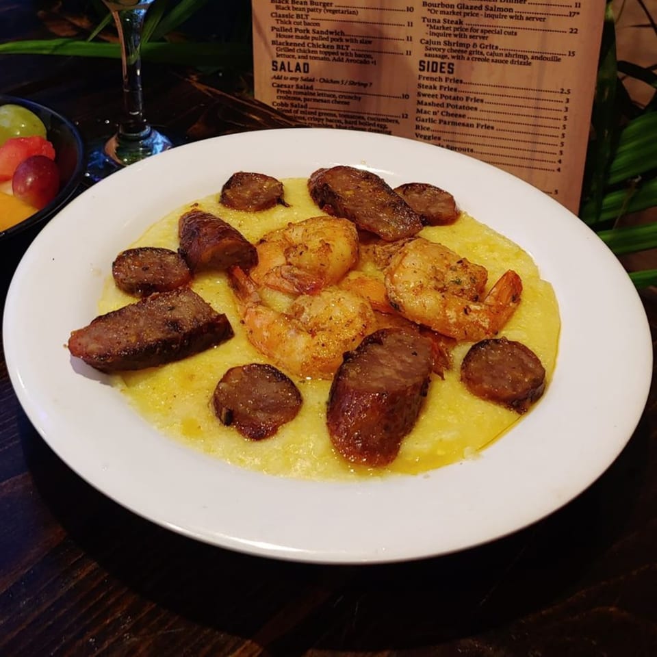 Cajun Shrimp & Grits with andouille sausage on a white plate, with a menu in the background.