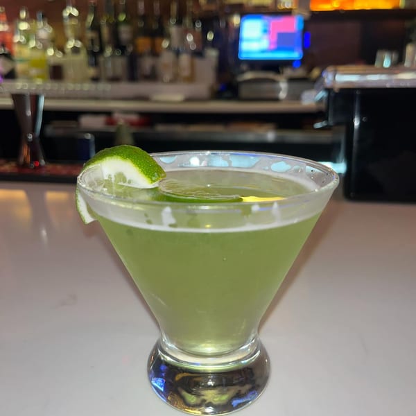Green martini with a lime wedge garnish on a white bar counter with blurred bottles in the background.