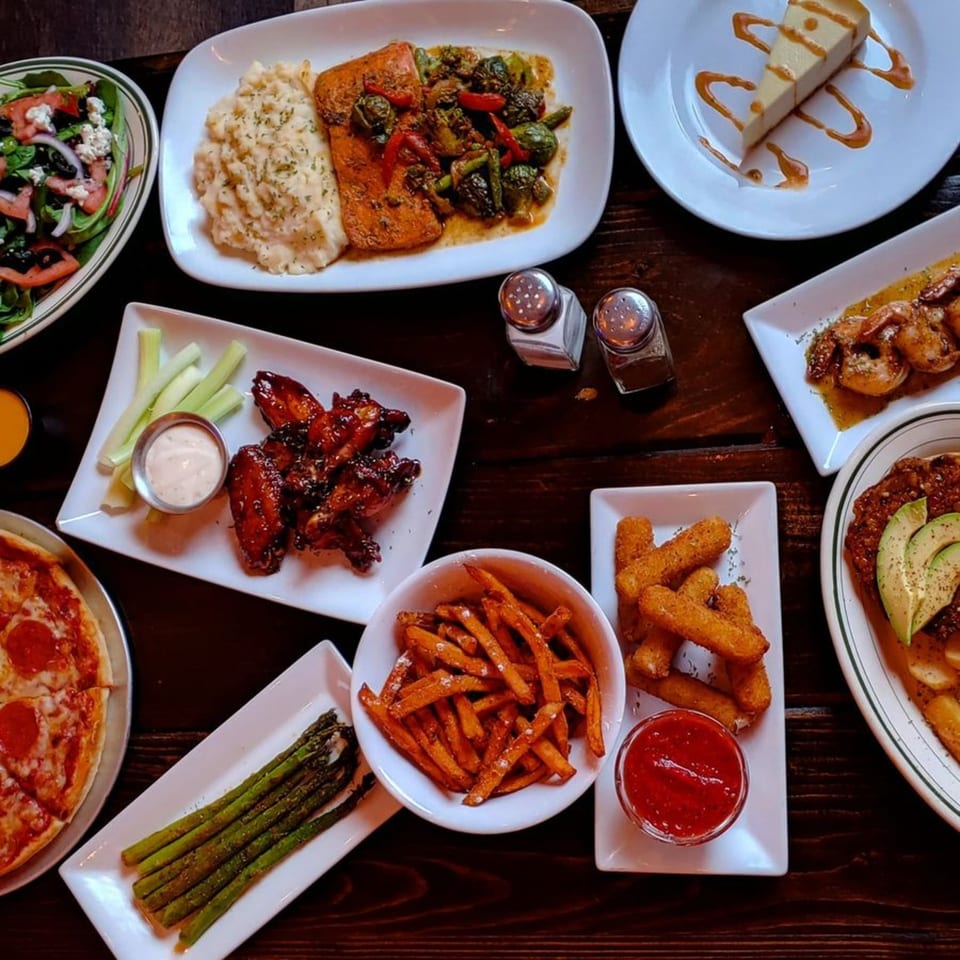 A feast of various dishes including salmon, wings, pizza, fries, and cheesecake on a dark wooden table.