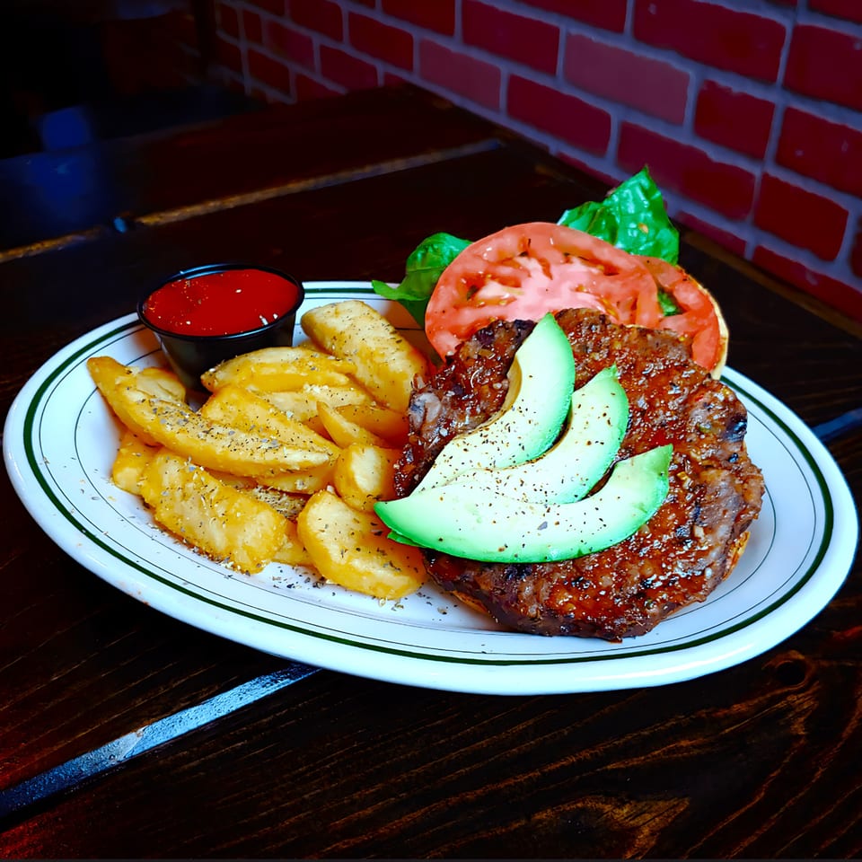 A burger with avocado slices, tomato, and lettuce, served with seasoned potato wedges and ketchup.