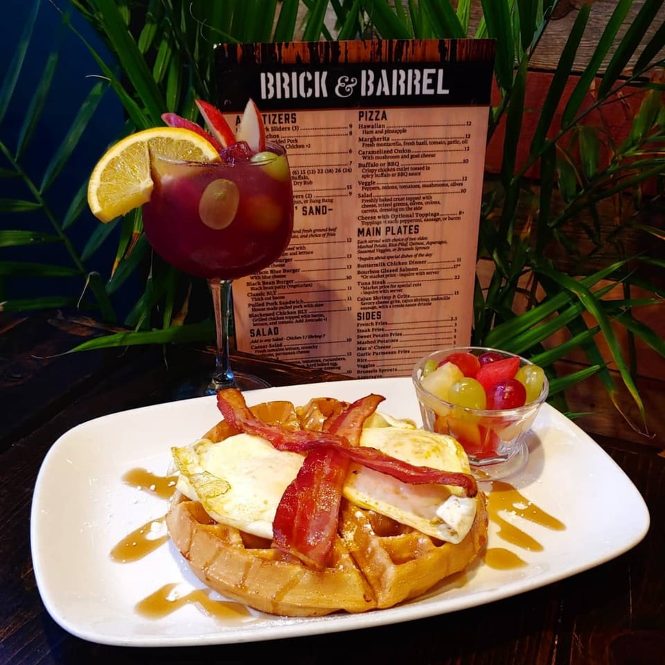 A waffle topped with fried eggs, bacon, syrup, a fruit cup, and a cocktail with a menu in the background.
