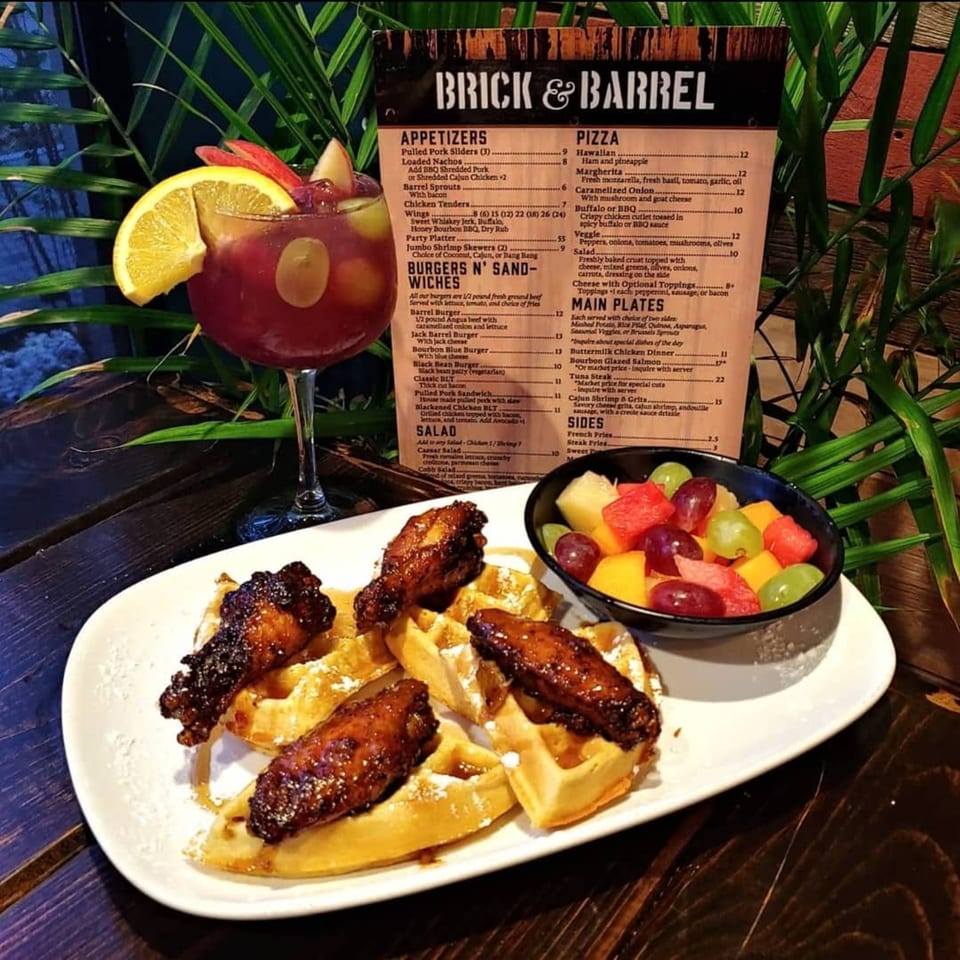Chicken and waffles with fruit salad and a cocktail, with a Brick & Barrel menu in the background.