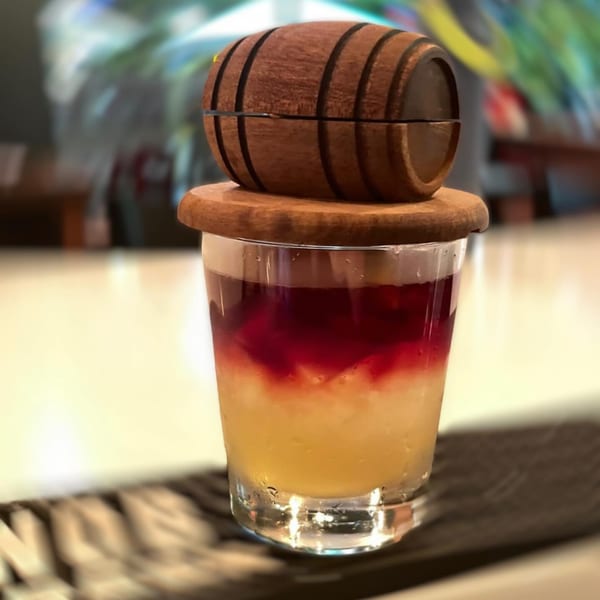 Layered cocktail with yellow and red liquids, topped with a small wooden barrel.