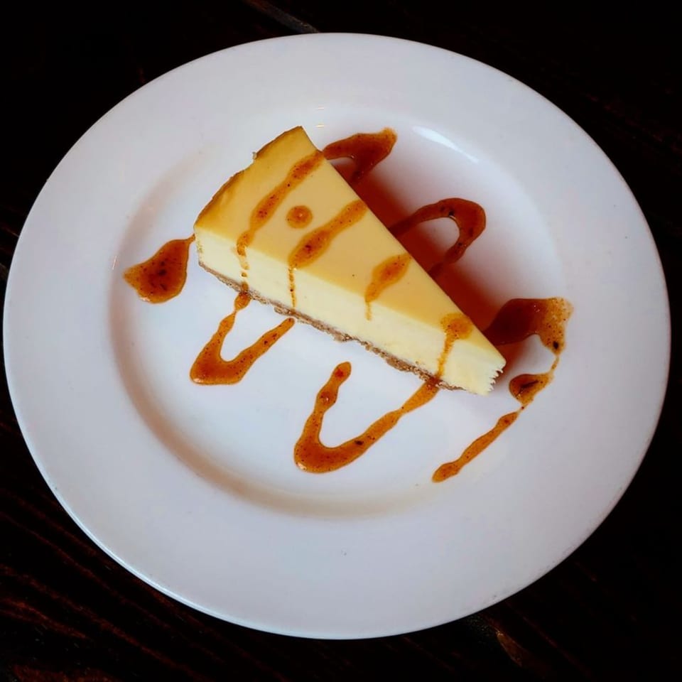 A slice of cheesecake with a graham cracker crust, drizzled with caramel sauce on a white plate.