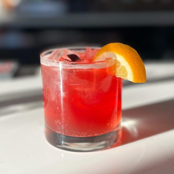 A vibrant red cocktail with ice and an orange slice garnish sits on a white surface.