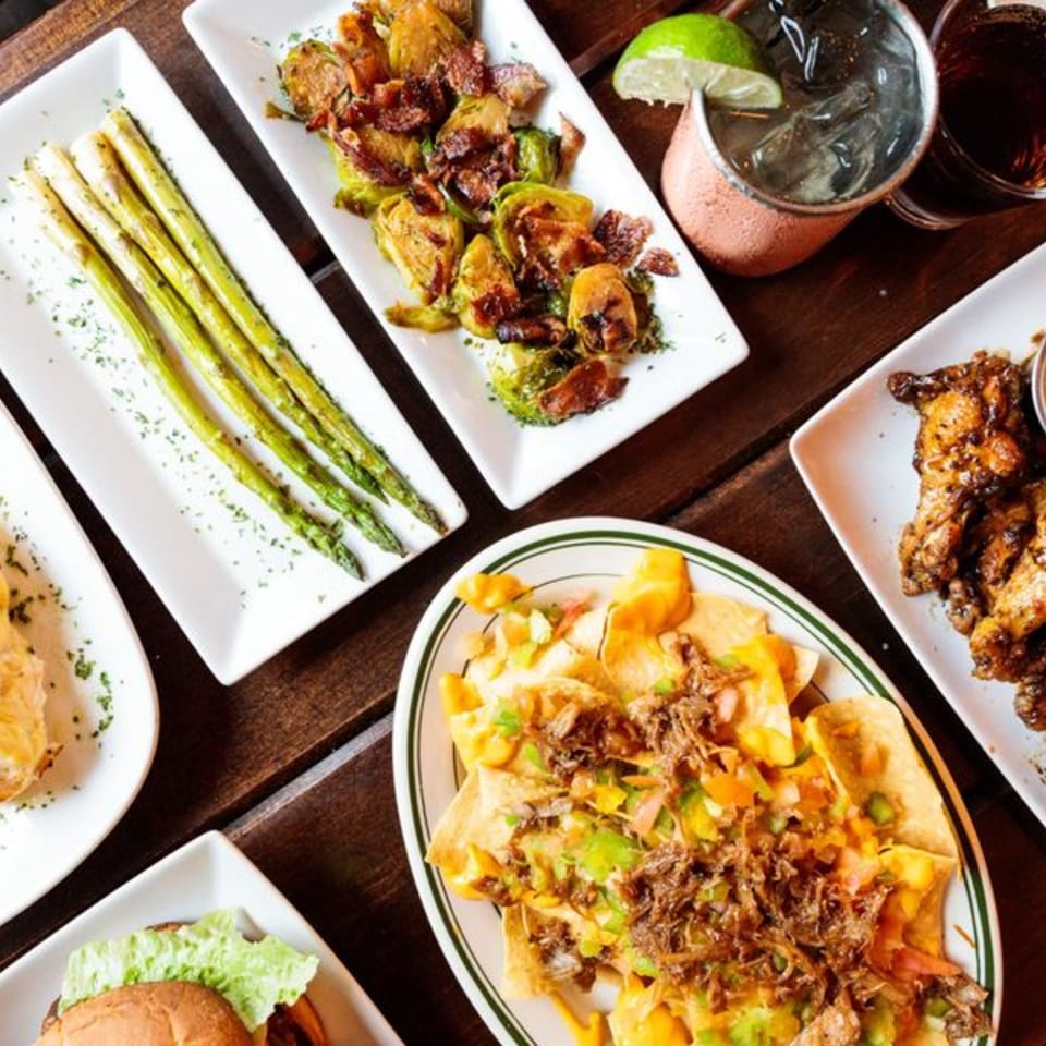 Table spread with nachos, asparagus, brussels sprouts, chicken wings, and drinks.