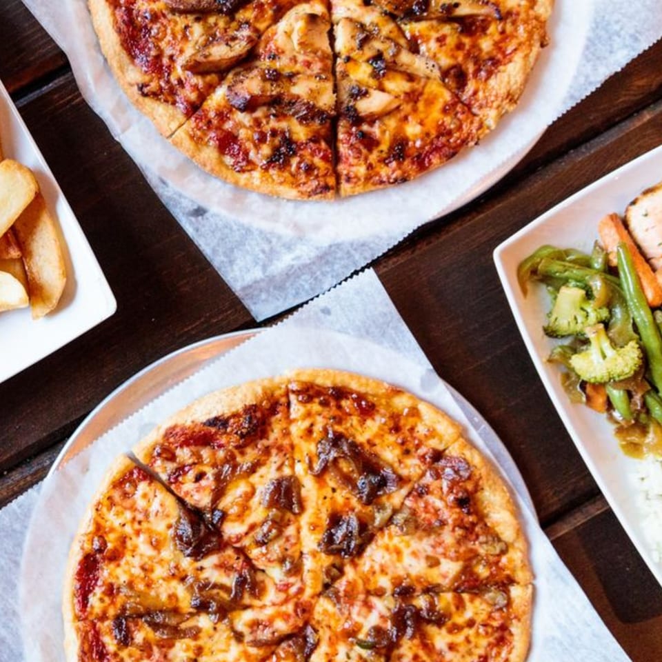 Two pizzas, a side of fries, and a plate of vegetables with chicken on a wooden table.