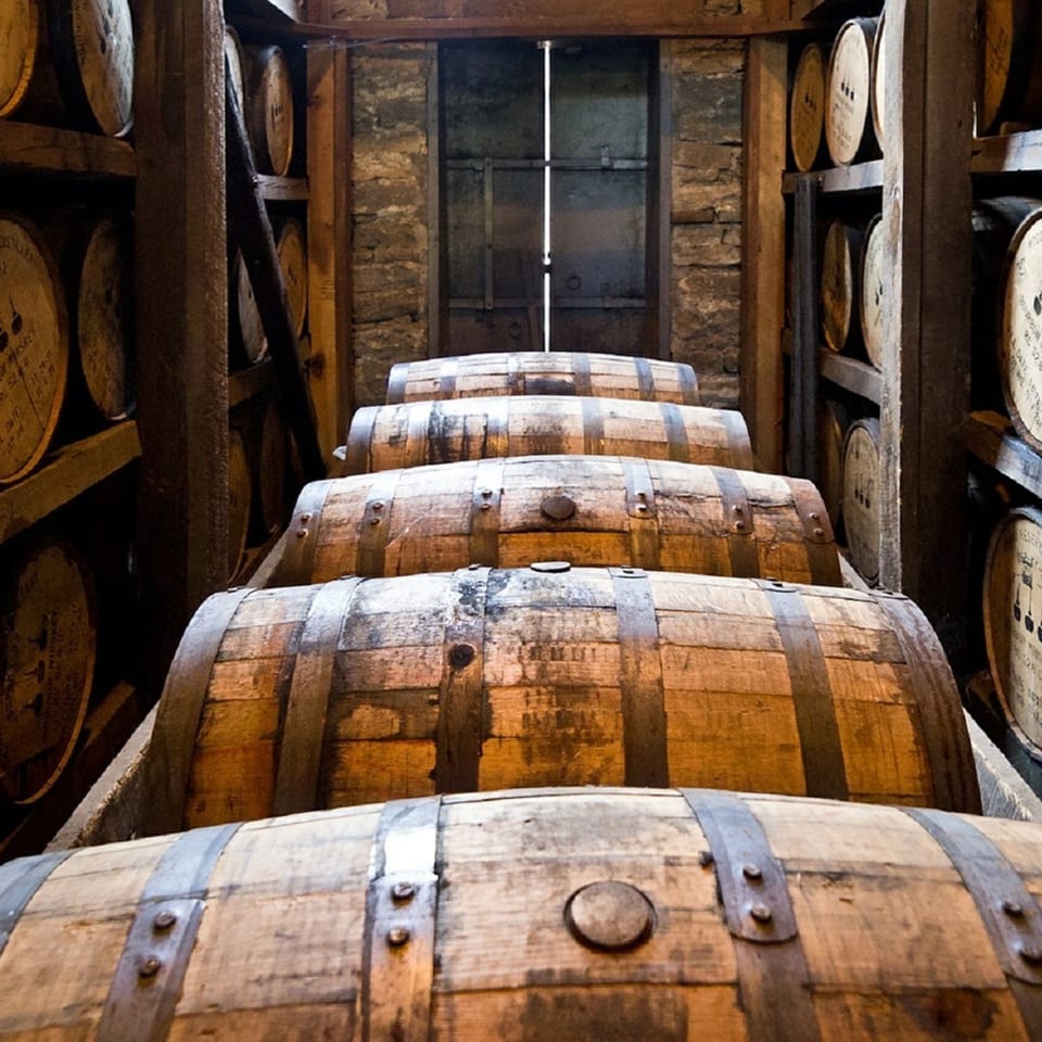 A long row of wooden whiskey barrels in a dimly lit cellar, with more barrels stacked on shelves.