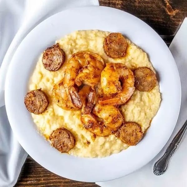 Shrimp and grits with sliced sausage on a white plate, next to a white napkin and fork.