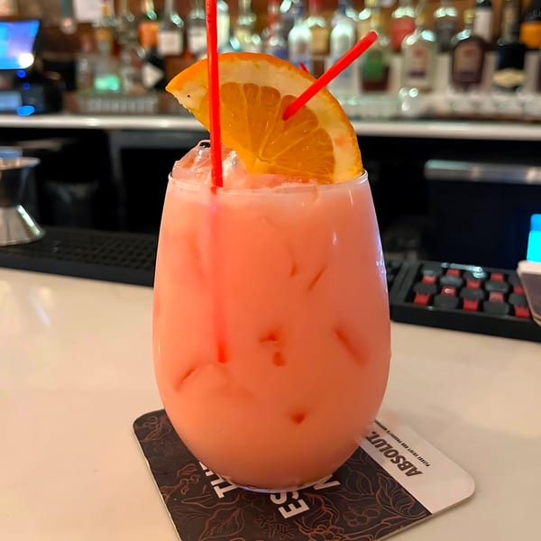 Pink cocktail with ice, an orange slice, and two red straws on a bar counter.